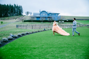 A bride wearing an Orange dress being chased by a groom wearing grey suit on a the green grass at Abbey Road Farms, Carlton, Oregon