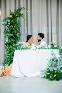 Couple enjoying the sweet moment at the sweetheart table decorated with greenery and pop of orange flowers located in the main reception hall of Abbey Road Farms in Carlton, Oregon