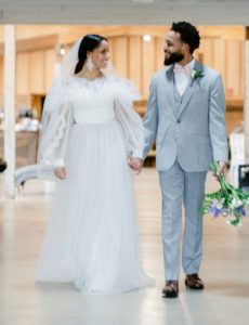 Bride wearing their traditional Alon Lavne White dress and a groom wearing a grey suit walking hand in hand inside Abbey Road Farms main hall of the reception