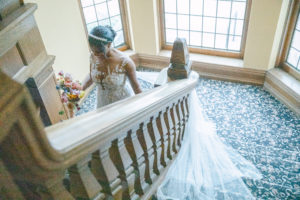 The bride wearing a gorgeous gown holding a bouquet walking down the stairways with her train wraps around the stairs and heading by the large bay window 