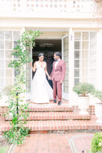 Bride wearing v-neck Justin Alexander wedding dress with tiara and Groom wearing burgandy suit from the Black Tux, holding hands by the door steps of Manor