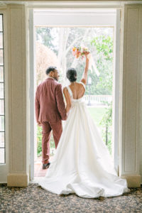 Groom wearing burgundy suit and bride wearing long detachable Justin Alexander dress with hand raise high holding a bouquet of flower in doorway 