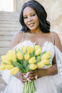 Black bride with hair down, wearing Pronovias gowns cold shoulder lace holding a bunch of yellow tulips. Makeup is a Timeless and elegant natural looking makeup